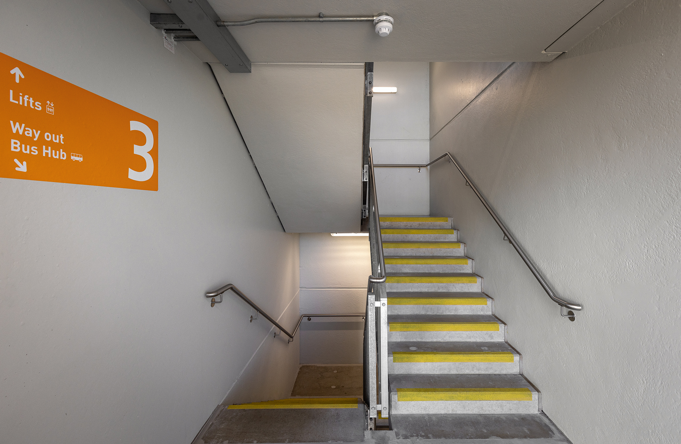 Helles Treppenhaus in einem Parkhaus auf Ebene 3, mit grauen Wänden, Geländern auf beiden Seiten und Stufen mit gelben Sicherheitskanten. An der Wand zeigt ein orangefarbenes Schild die Richtung zu Aufzügen, Ausgang und Bus-Hub.