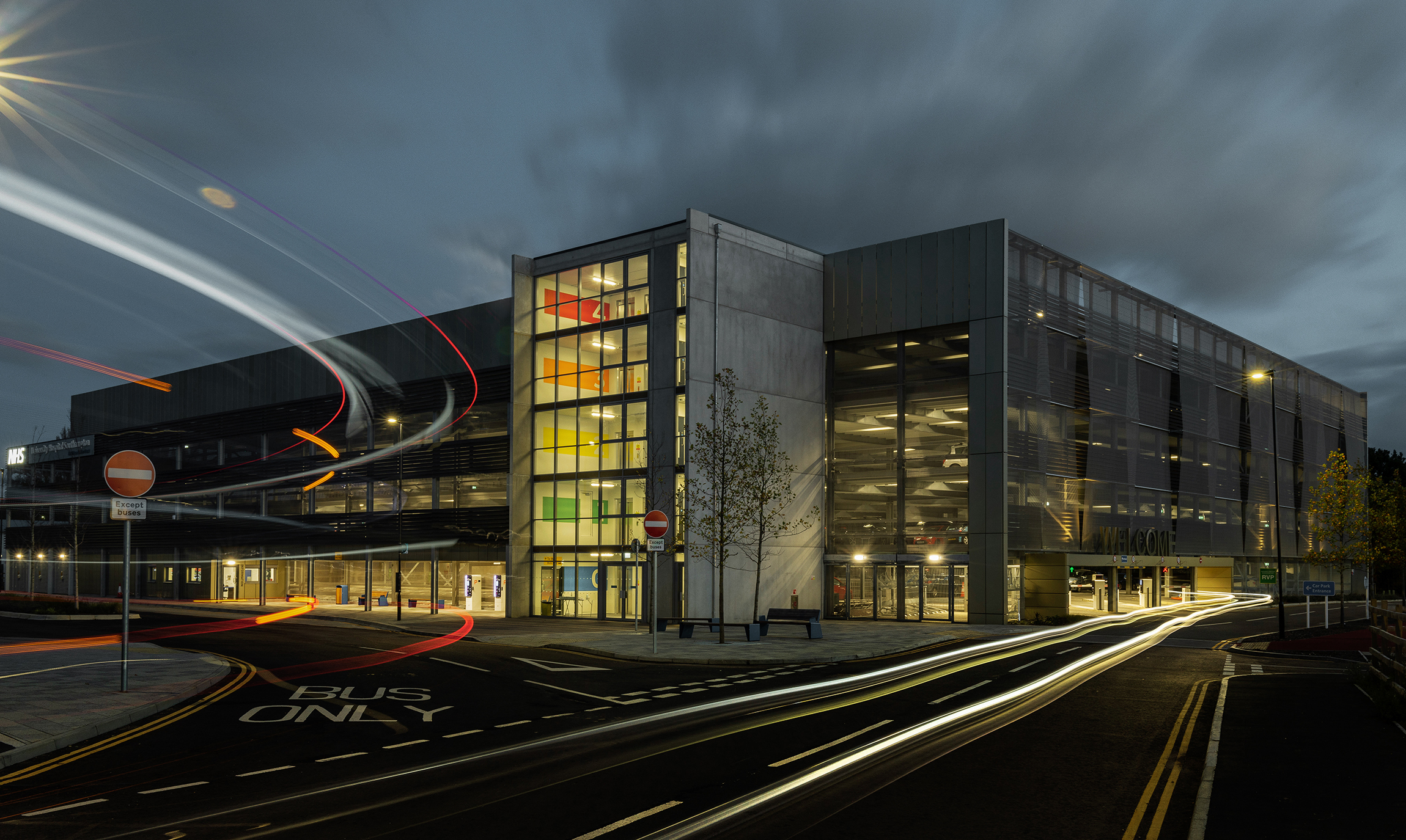Beleuchtetes Parkhaus bei Nacht mit farbigem Treppenhaus und Lichtspuren vorbeifahrender Autos.