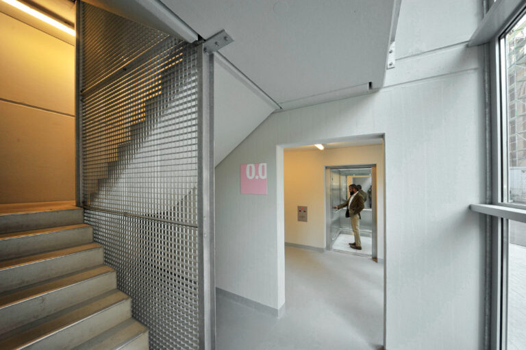 Treppenhaus mit Aufzug im Parkhaus, Betonstufen, Gittertrennwand und Geschosskennzeichnung.