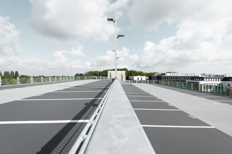 Zentrale Perspektive über das Parkdeck mit Beleuchtungsmast, Parkplatzmarkierungen und Stadtbebauung im Hintergrund.