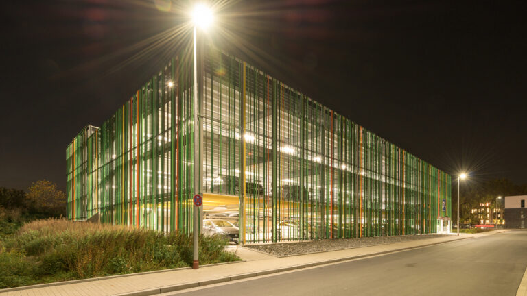 Nächtliche Außenansicht eines mehrgeschossigen Parkhauses mit grüner Lamellenfassade und heller Innenbeleuchtung an einer ruhigen Straße.