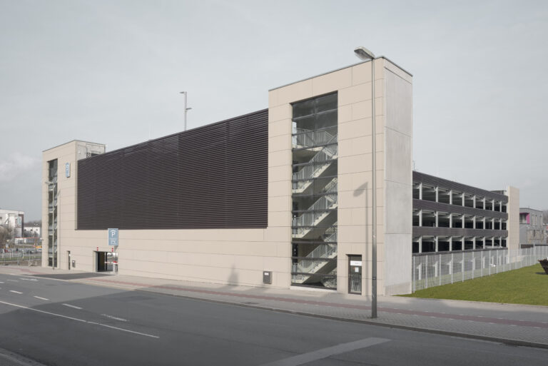 Schrägansicht des Parkhauses mit Zufahrt, Glas-Treppenhaus und umliegender Straßen- und Wohnbebauung.