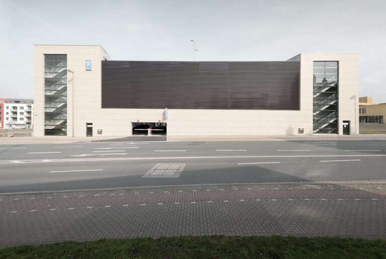 Moderne Parkhausfassade mit zentraler Zufahrt und seitlichen Treppenhäusern aus Glas an einer breiten Straße.