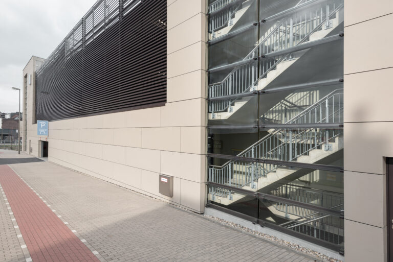 eitenansicht der Parkhausfassade mit Glas-Treppenhaus und dunkler Lamellenverkleidung entlang des Gehwegs.