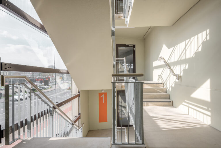 Helles Treppenhaus im Parkhaus mit Glasfassade, Edelstahlgeländern und Blick auf umliegende Geschäfte.