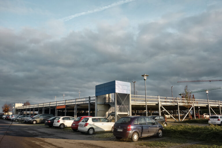 Parkplatz mit abgestellten Autos vor einem zweigeschossigen Parkdeck mit Treppenaufgang.