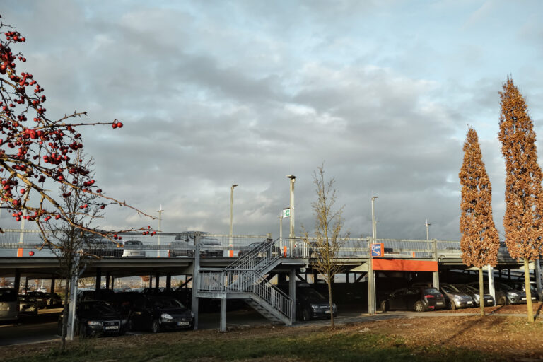 Offenes Parkdeck mit zwei Ebenen, Außentreppe und parkenden Autos unter bewölktem Himmel.