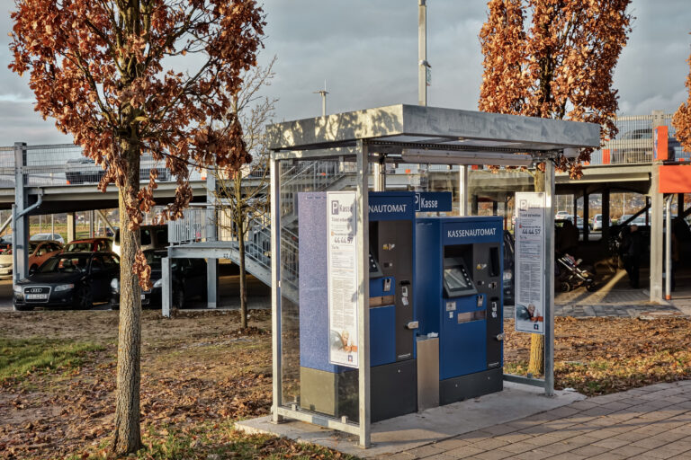 Kassenautomaten unter einem kleinen Unterstand vor einem Parkdeck mit Treppe im Hintergrund.