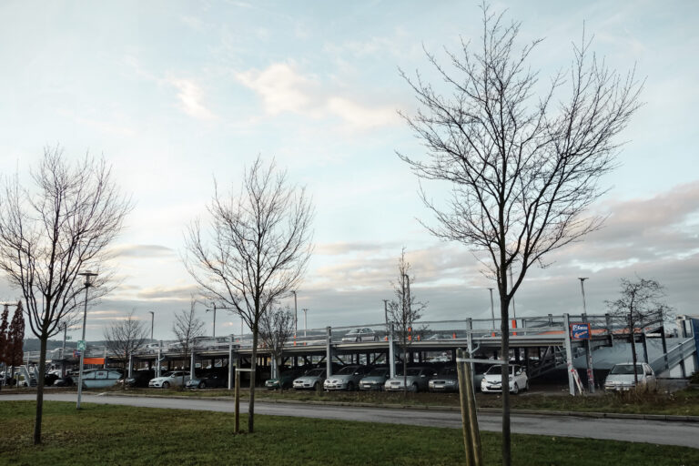 Langgestrecktes Parkdeck mit parkenden Autos, umgeben von Bäumen und Grünfläche.