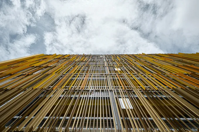 Sicht auf die gelbe Lamellenfassade eines Parkhauses mit rhythmisch angeordneten Metallelementen vor leicht bewölktem Himmel.