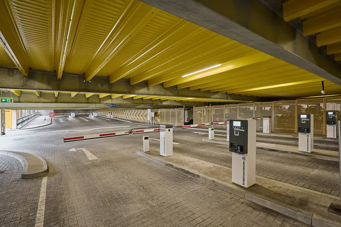 Einfahrtsbereich eines Parkhauses mit Schrankenanlage und Bezahlautomaten unter gelber Stahlträgerdecke, modern und übersichtlich gestaltet.