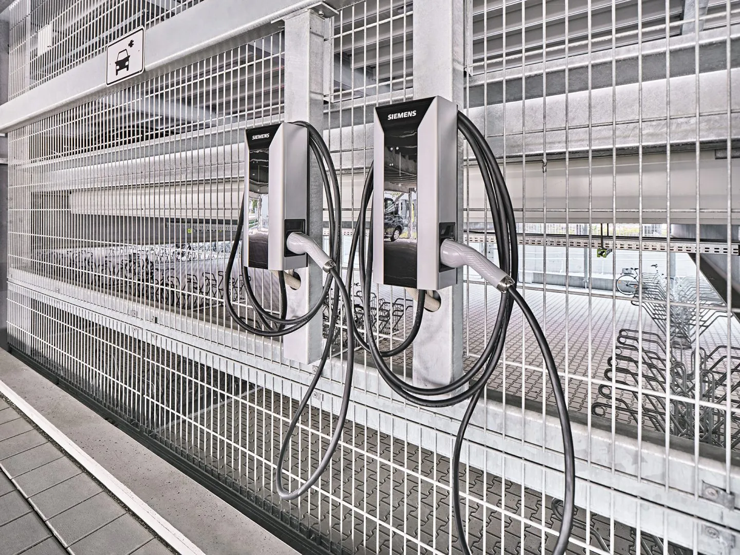 „Zwei Ladestationen für Elektrofahrzeuge im Parkhaus, montiert an silbergrauer Gitterfassade mit angeschlossenen Ladekabeln.