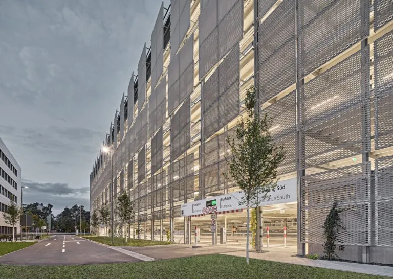 Abendliche Straßenansicht eines mehrgeschossigen Parkhauses mit silbergrauer Metallfassade, beleuchteter Einfahrt ‚Eingang Süd‘ und Baumreihe entlang der Zufahrt.