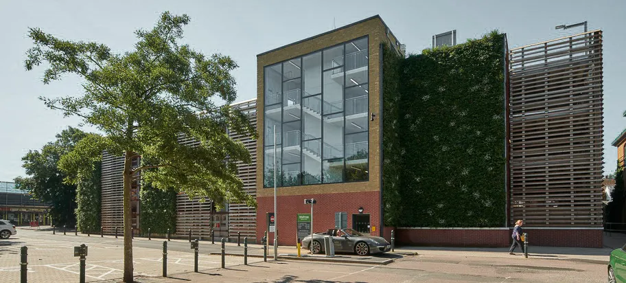 Modernes Parkhaus mit großer Glasfront des Treppenhauses und begrünter Fassade, umgeben von Bäumen und Gehwegen.