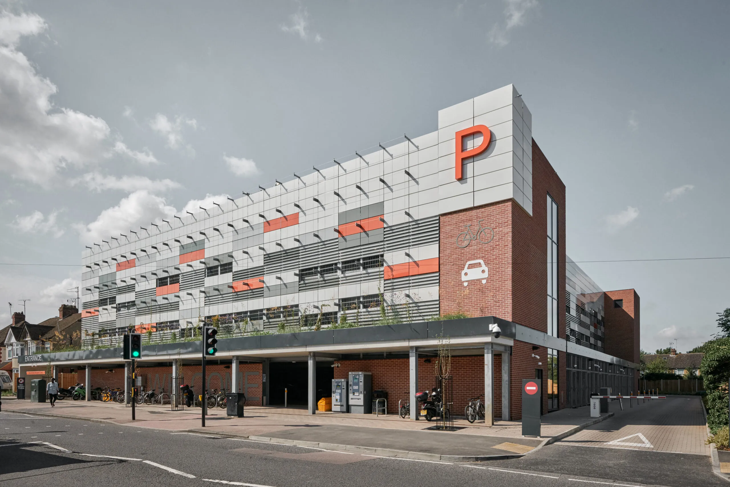 Schrägansicht eines modernen Parkhauses mit heller Paneelfassade, orangefarbenem „P“ und Fahrrad-/Auto-Symbolen.